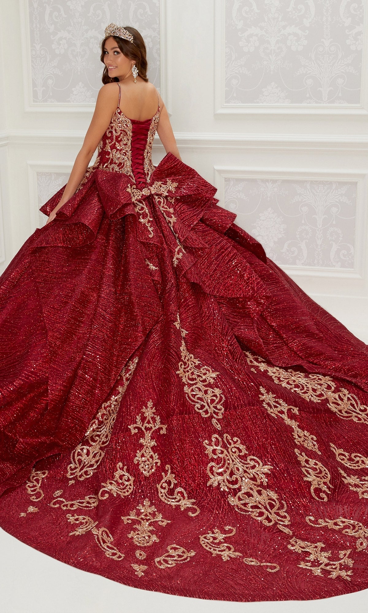 A woman in the PR22142 By Princesa Quinceanera Dress—red with gold embroidery and detachable sleeves—stands in a white room, facing away and looking over her shoulder.